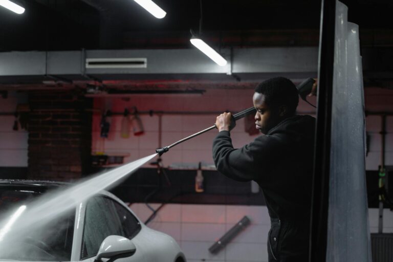 A man uses a high-pressure hose to wash a car in an indoor garage setting.